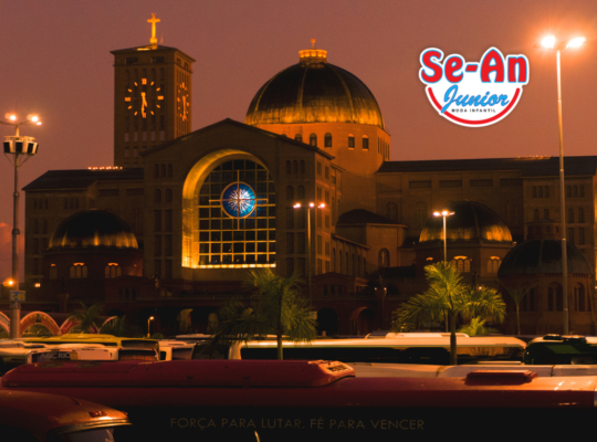 Na imagem há a Catedral principal de Aparecida / São Paulo. A foto foi tirada à noite. No canto superior direito, há a logomarca da Se-An Junior moda infantil.