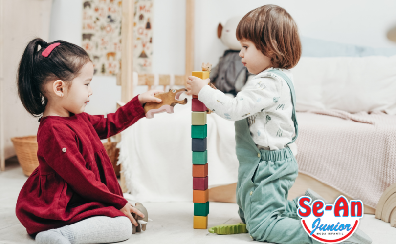 A imagem pretende ilustrar o post sobre a melhor roupa infantil. Nela há duas crianças brincandoem um quarto. A cama, com lençol e edredom claros, está aofundo. A menina está de ladopara a câmera, voltada para a direita, de frente para o menino. Ela está com o cabelo preso em um rabo de cavalo,usa um vestido bordô de manga comprida e meias-calça brancas. Não está usando sapatos. O menino veste um macacãojeans com blusa de manga comprida branca e meias brancas. Albos estão empilhando blocos coloridos.