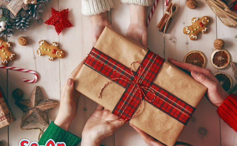 A imagem ilustra o artigo sobre presentes de Natal. É uma foto tirada a partir de uma visão superior (de cima). A imagem mostra três pares de mãos segurando um presente em cima de uma mesa de madeira branca. Cada par de mãos está vestindo uma blusa de lã de cor diferente: branca, vermelha e verde. O presente está embrulhado em papel kraft e com uma fita avermelhada. Na mesa há objetos que remetem ao Natal como parte de uma vesta de vime, guirlanda, bolinhas, estrelinhas, boneco de gengibre e outros.