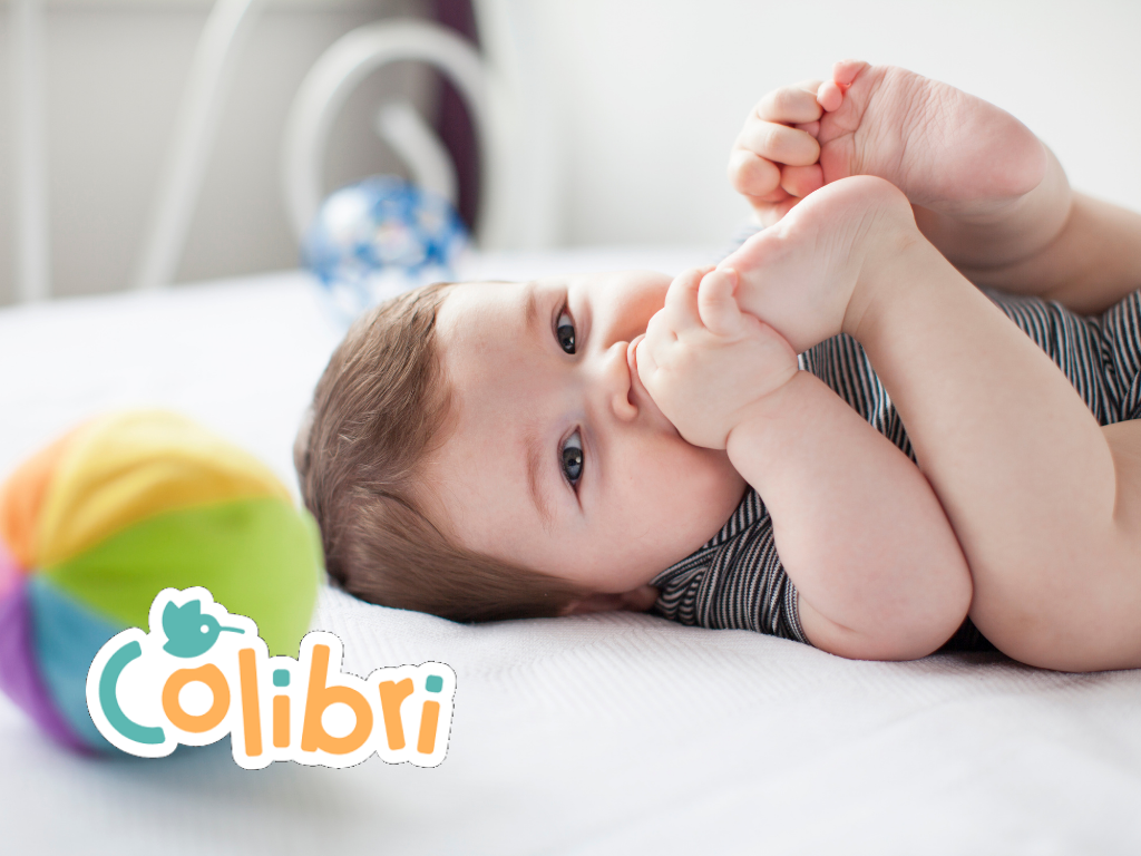 A imagem pretende ilustrar o post sobre os enxovais para bebê da Colibri. Ela apresenta um bebê, deitado na cama, com pernas para cima. Com uma das mãos ele segura um dos pezinhos e o leva à boca. Em primeiro plano há uma bola, desfocada.