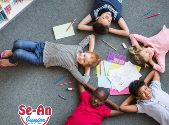 A imagem que ilustra o artigo sobre roupa infantil barata mostra5 crianças deitadas em um chão com carpete cinza. Há várias folhas de papel e lápis espalhados pelo chão. As crianças estão formando um círculo com as pernas para fora, as mãos atrás da cabeça e os cotovelos tocando-se.