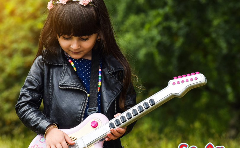 A imagem que ilustra o artigo sobre moda infantil feminina estilosa tem o foco em uma garotinha com uma guitarra cor de rosa. Ao fundo há árvores e arbustos. Ela tem cabelo escuro e franja. em seu cabelo há uma tiara de flores e ela veste um vestido azul com uma jaqueta que parece couro na cor preta. Ela está segurando a guitarra como se a estivesse tocando.