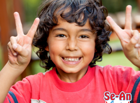 A imagem que ilustra o texto sobre roupa infantil masculina estilosa mostra um menino com cabelos encaracolados. Ele está sorrindo para a câmera e com os dedinhos das mãos levantadas formando o V da vitória. Ele usa uma camiseta branca, vermelha e azul.