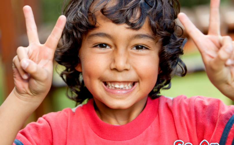 A imagem que ilustra o texto sobre roupa infantil masculina estilosa mostra um menino com cabelos encaracolados. Ele está sorrindo para a câmera e com os dedinhos das mãos levantadas formando o V da vitória. Ele usa uma camiseta branca, vermelha e azul.