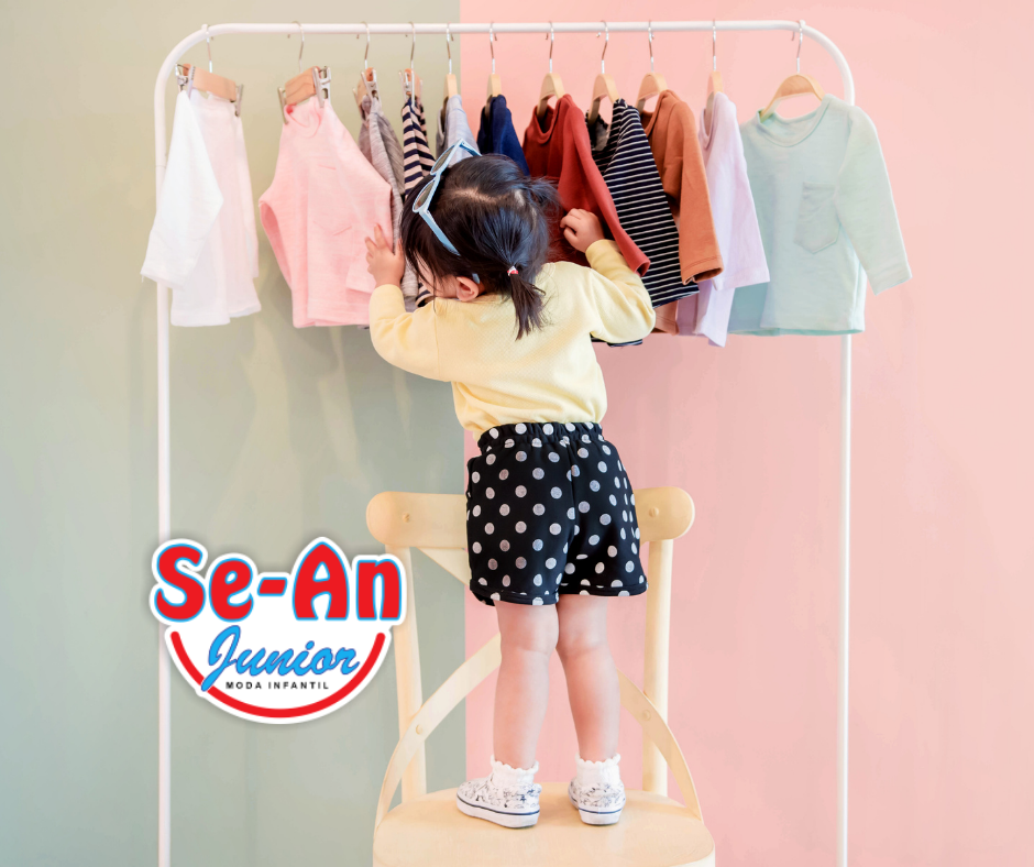 A imagem que ilustra o artigo sobre roupa infantil de marca mostra uma menina em cima de uma cadeira. Ela está mexendo em uma arara de roupas infantis. Há roupas de várias cores. A arara é branca e a parede ao fundo metade rosa e metade cinza. A menina está de costas para a câmera. Ela tem cabelos pretos, pele branca. Seus cabelos estão amarrados em dois rabinhos de cavalo e ela tem um óculos de sol na cabeça. Ela veste uma blusa de manga amarela, um short preto de bolinhas brancas e tênis brancos com alguns desenhos coloridos e meias brancas.
